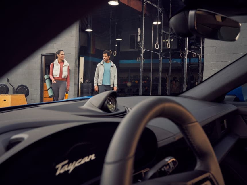 View from inside a Porsche Taycan, showing a man and woman walking outside a gym, with gym equipment visible in the background.